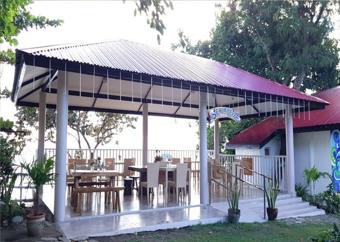 Covered outdoor dining area with tables and chairs near a building, water in the background.