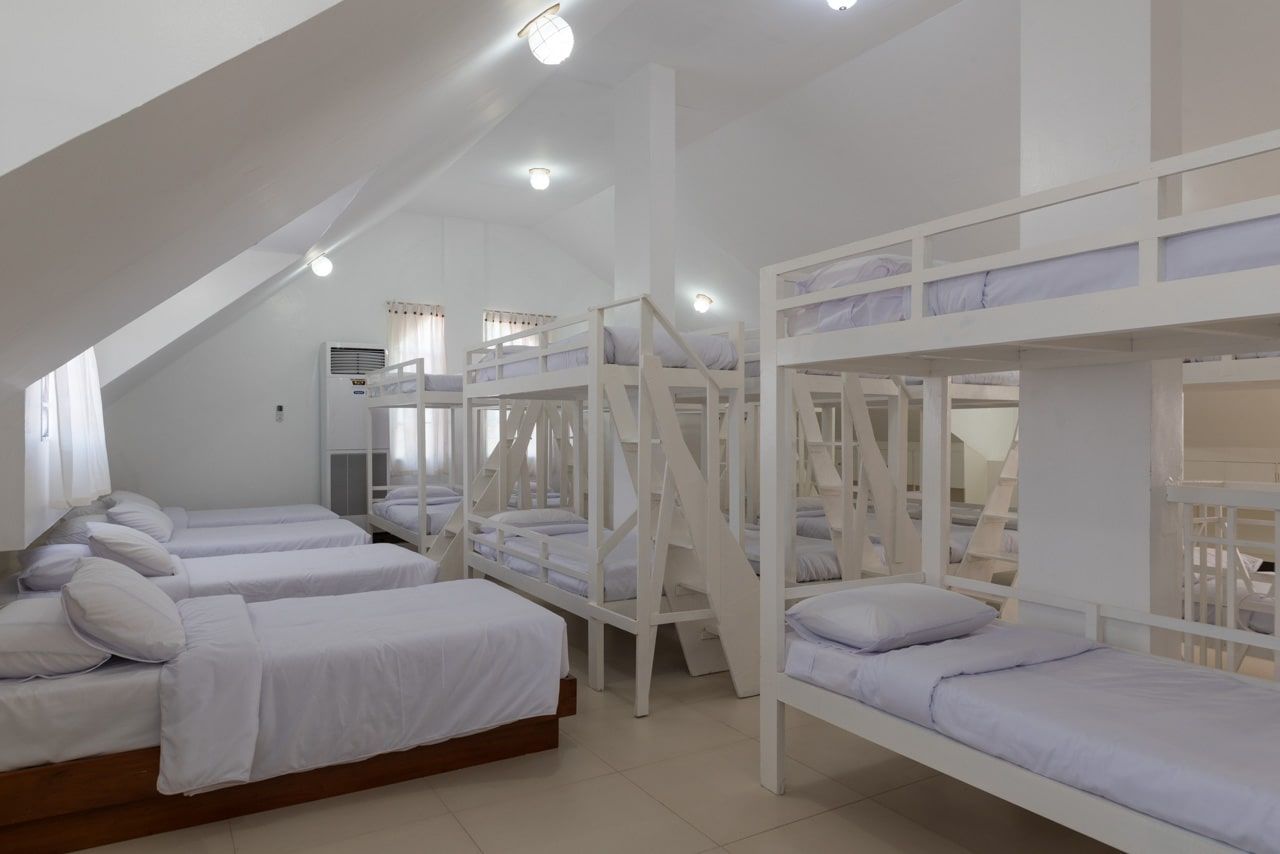 Bright, white-walled dorm room with multiple bunk beds and single beds; windows and recessed lighting.