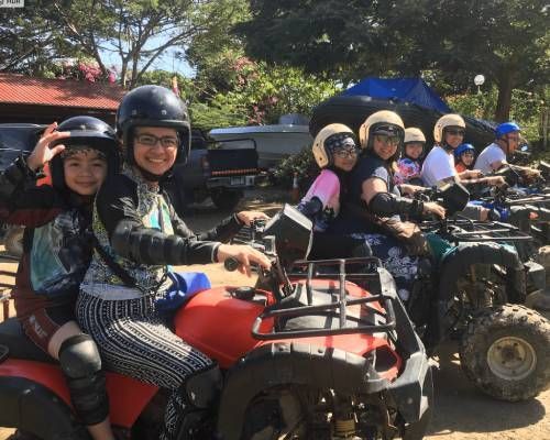 People on ATVs, wearing helmets and safety gear, in a sunny outdoor setting.