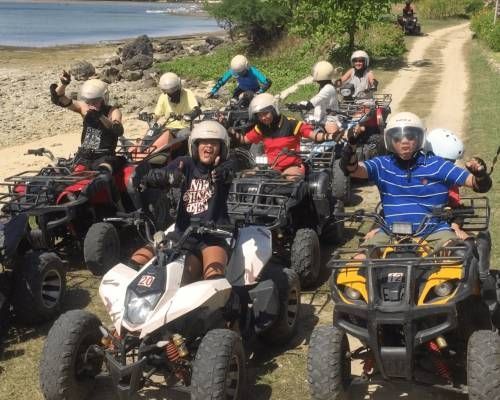 Group of people on ATVs, wearing helmets, riding on a dirt path near a body of water, waving.