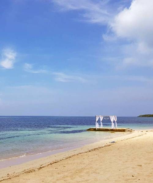 Beach scene with clear blue sky, ocean, sandy shore, and white draped gazebo on a small pier.