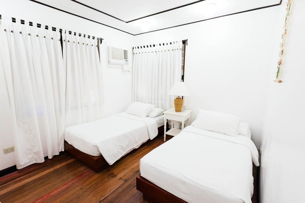 Bright white bedroom with two twin beds, white curtains, and a nightstand with a lamp.