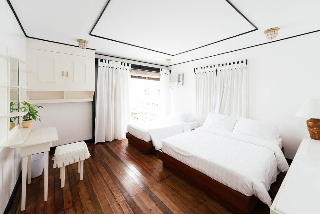 Bright bedroom with two beds, white curtains, and wood flooring. A small table with a chair is in the corner.