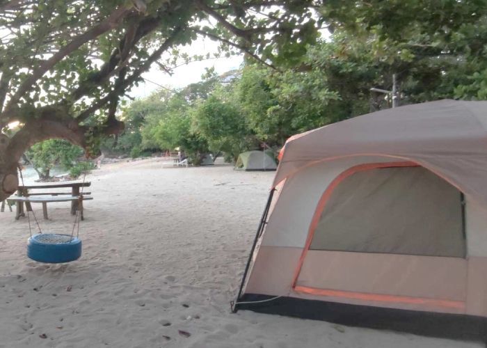 Tent on sandy beach near a tire swing and picnic table, trees in the background.