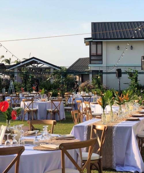 Outdoor event tables set for a gathering on a grassy lawn; houses and string lights in the background.