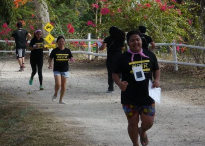 People running outdoors on a dirt path, wearing matching black shirts, a pink scarf, and colorful shorts.