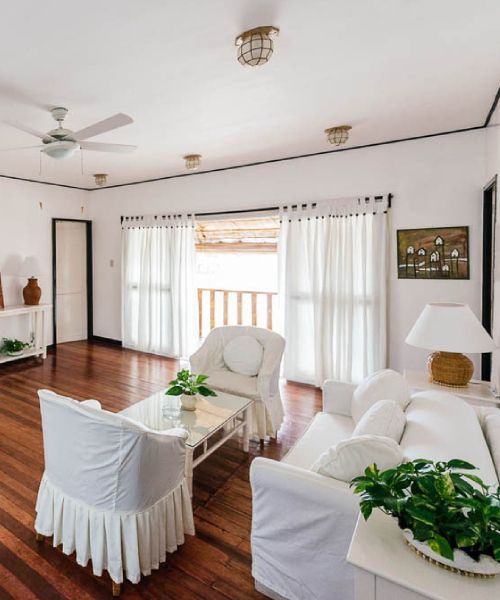 Bright living room with white furniture, wooden floors, and sheer white curtains.