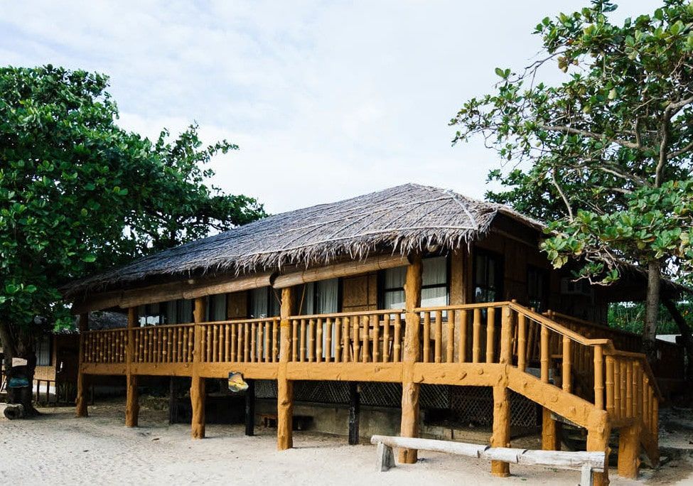 Wooden bungalow with thatched roof and deck on a sandy beach, under a cloudy sky.
