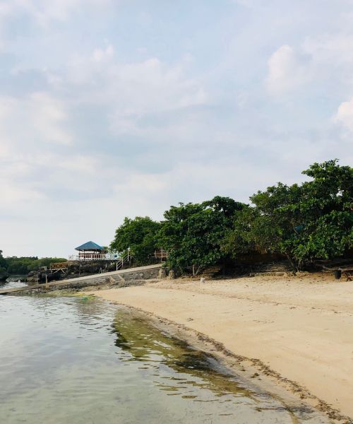 Sandy beach with clear water, trees, and a small structure under a cloudy sky.