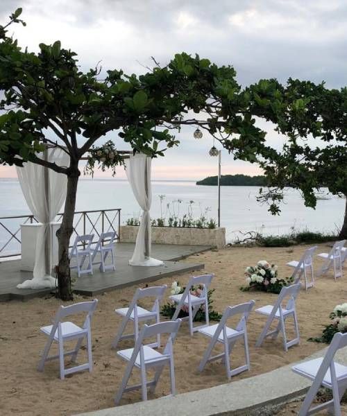 Wedding ceremony setup on a beach with white chairs, draped fabric, and flowers. Ocean and trees in the background.