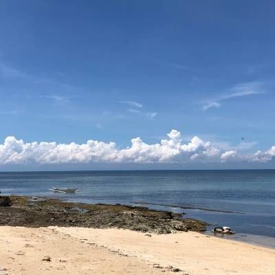 Beach scene: sandy shore, rocky edge, calm blue ocean, puffy white clouds, and bright blue sky.