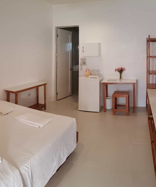 Small, minimalist hotel room with a bed, desk, mini-fridge, and open door to bathroom.