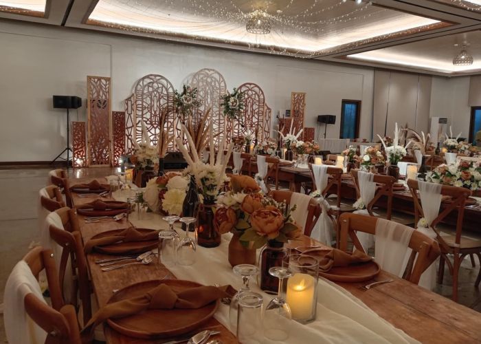 Wedding reception with long wooden tables, flower centerpieces, and decorative backdrop in a large hall.