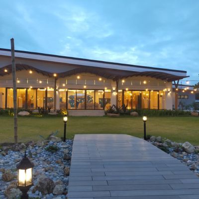 Wood walkway leading to a lit building at dusk, with string lights and garden lamps.