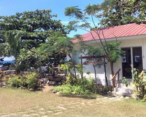 A white building with a red roof surrounded by green trees and foliage, near a grassy area.