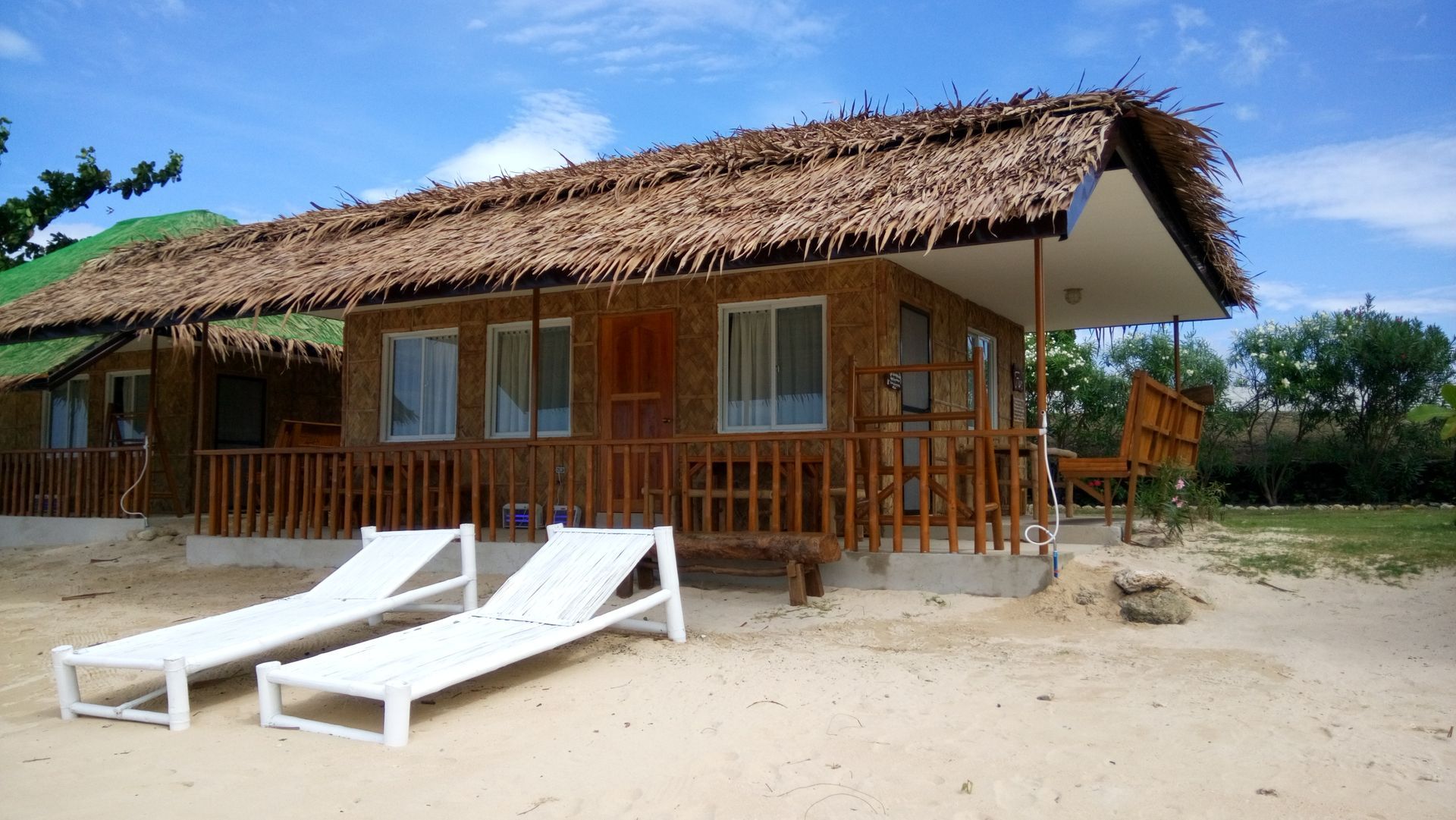 Beachside bungalows with thatched roofs and wooden railings, two white lounge chairs sit in front.