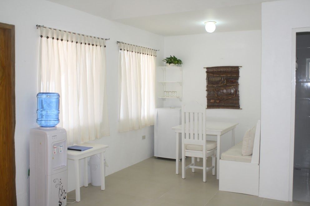 Small white dining area with water cooler, table, chairs, fridge, and window curtains.