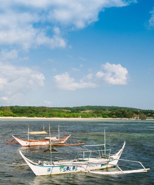 Two white boats with outriggers float on the sea near a sandy beach under a blue sky with clouds.