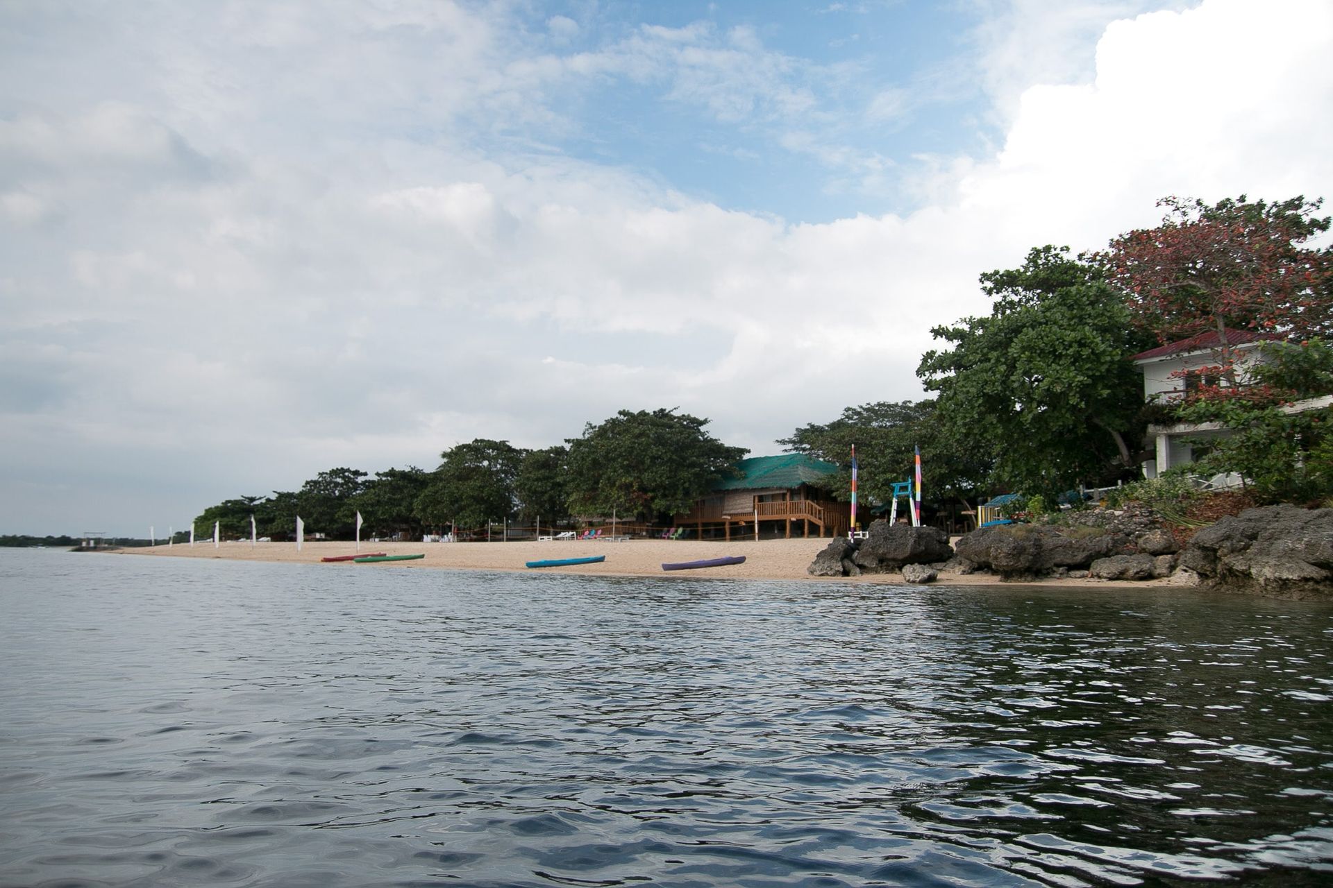 Calm water, sandy beach, buildings, and trees under a cloudy sky.