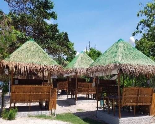 Several bamboo huts with thatched roofs on a beach, under a bright blue sky.
