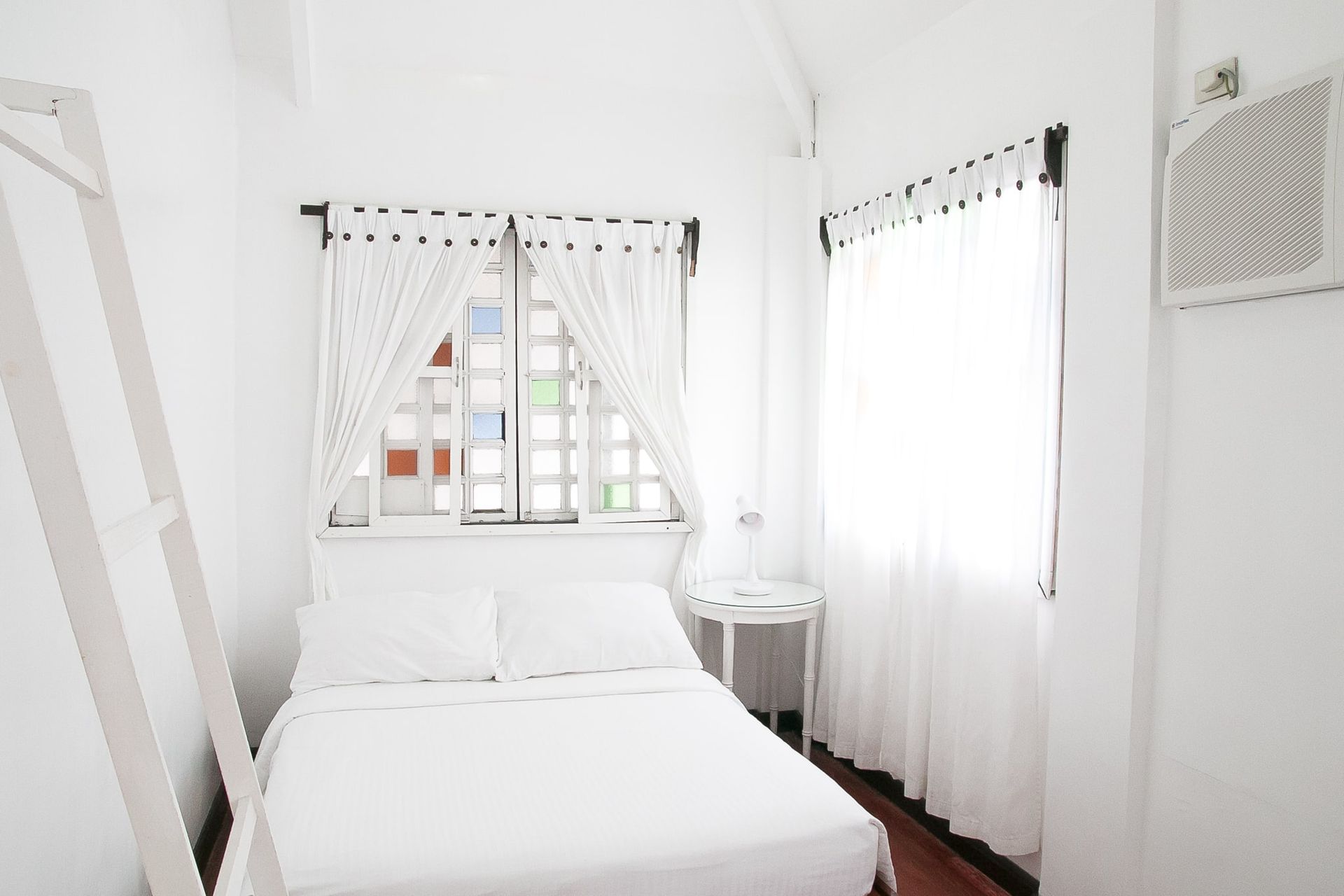 Bright white bedroom with bed, window, and curtains. A ladder leans against the wall.