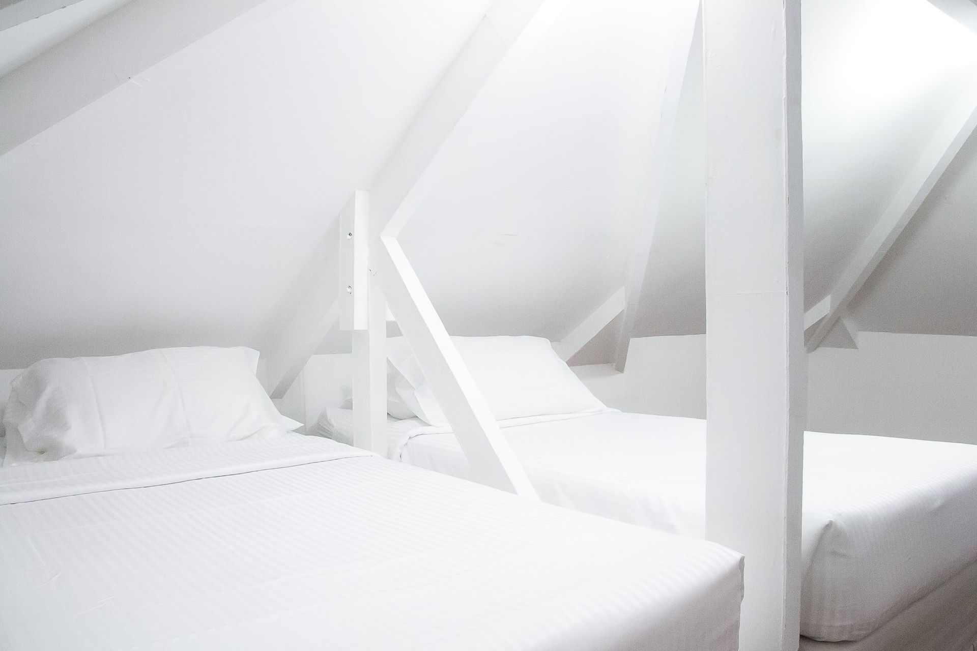 White attic bedroom with two beds. White beams support the slanted ceiling.