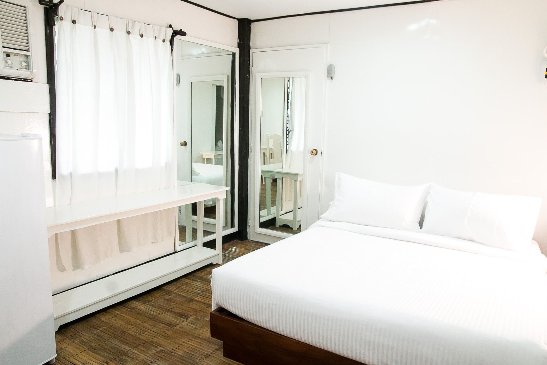 White bedroom with bed, desk, mirror, and window with curtains.