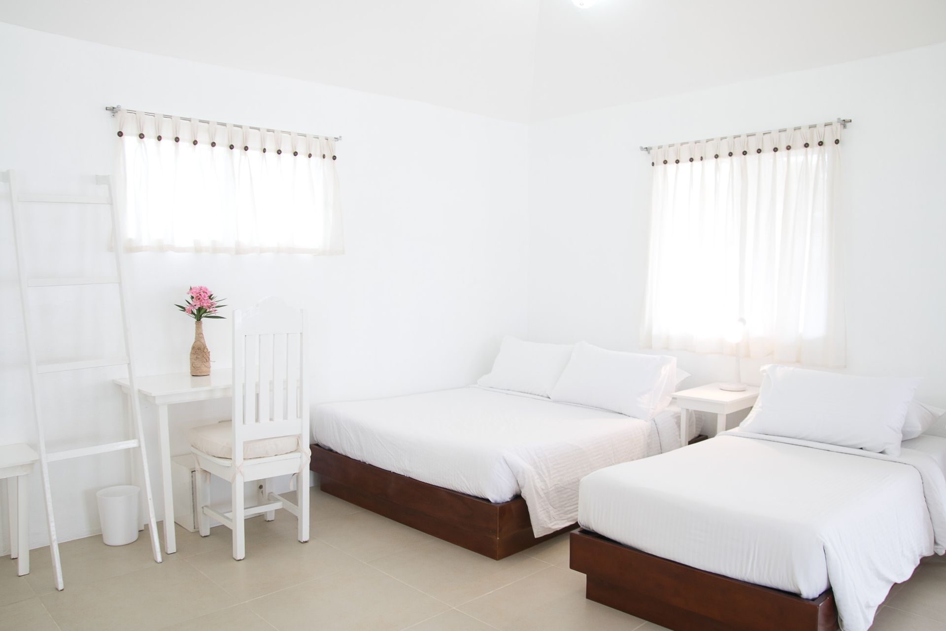 White bedroom with two beds, small desk, and two windows with sheer curtains.
