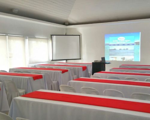 Conference room with tables covered in white linens and red runners, projector screen, and a projected image.