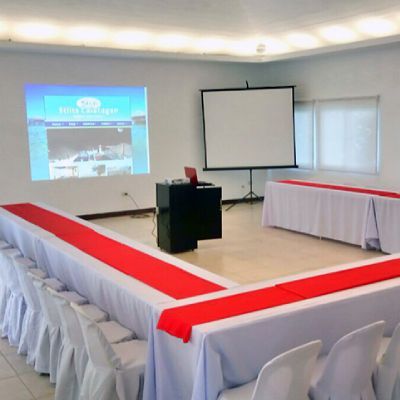 Conference room with U-shaped tables, white linens, red runners, projector screen, laptop, and chairs.