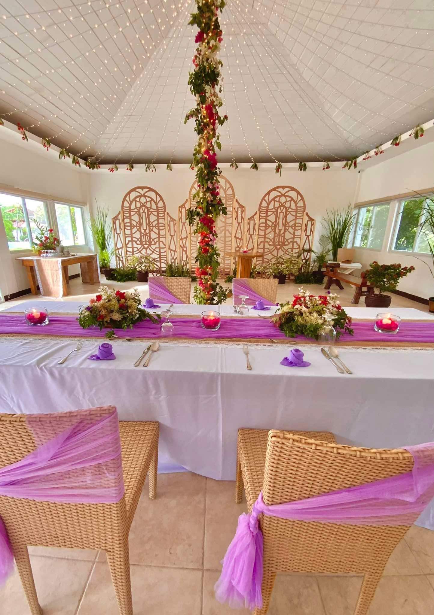Wedding reception table set with purple runner, floral centerpieces, and decorated wicker chairs.