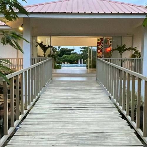 Wooden bridge leading to an open doorway with a pool view. White building, pink roof, and lush greenery.