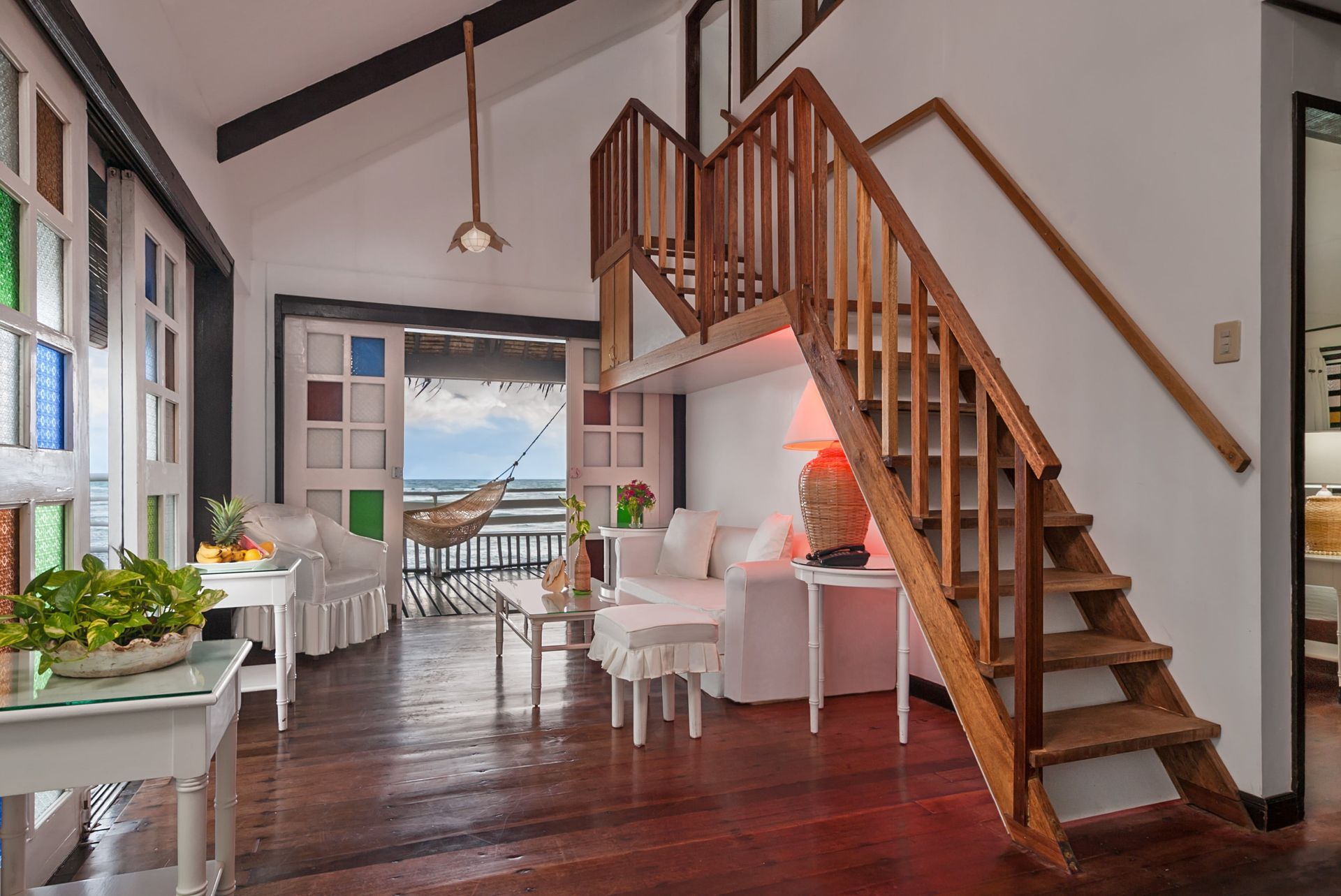 Interior of a bright beach house with a loft, wood stairs, white furniture, and a view of the ocean.