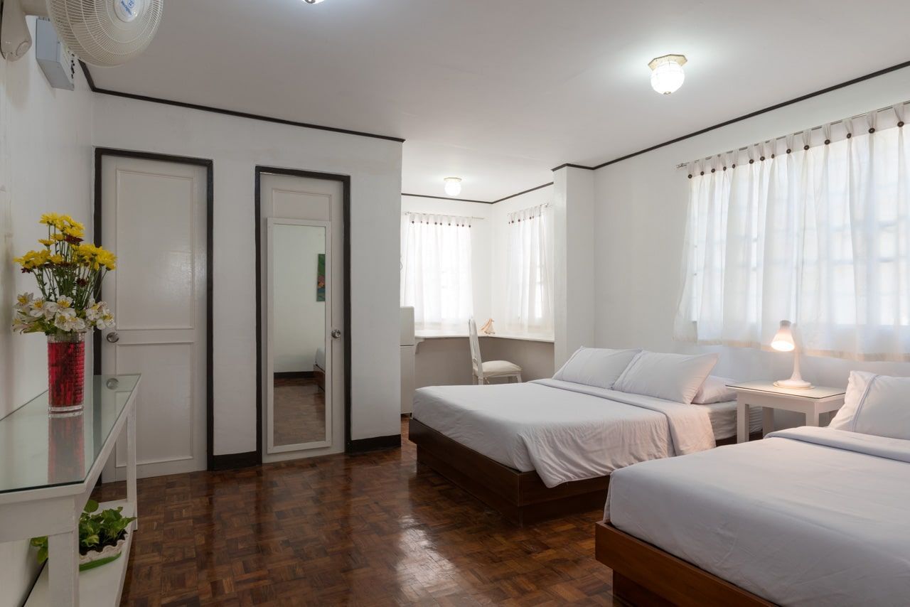 Bright hotel room with three beds, white walls, and wooden floors.