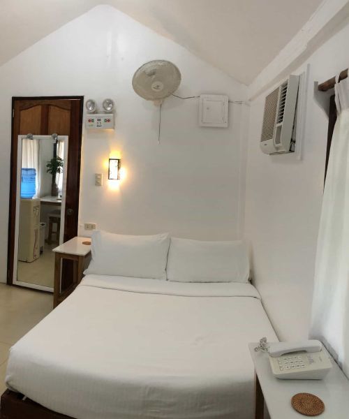 A white-walled hotel room with a bed, small side tables, a door, and an air conditioner.