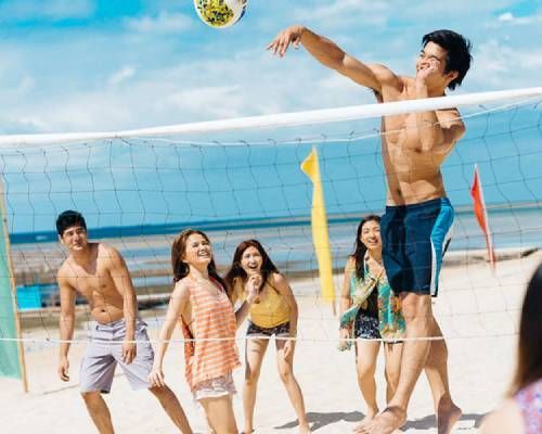 People playing beach volleyball; shirtless man spiking the ball, others watching, ocean background.