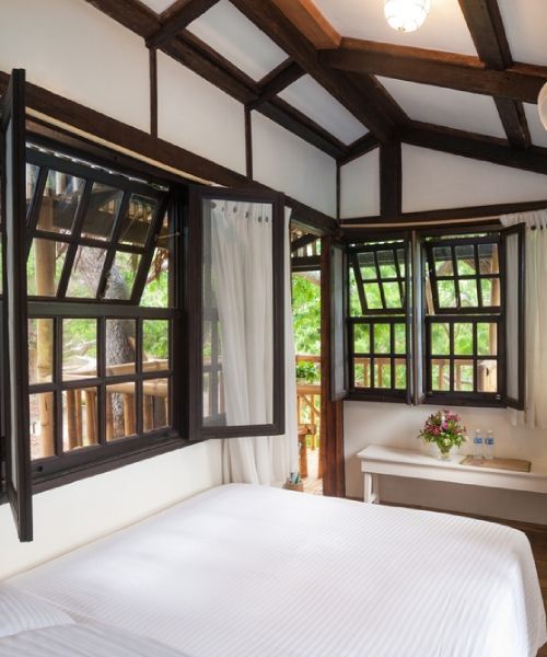 Bedroom with a white bed, open dark-framed windows, and a white ceiling with wooden beams.