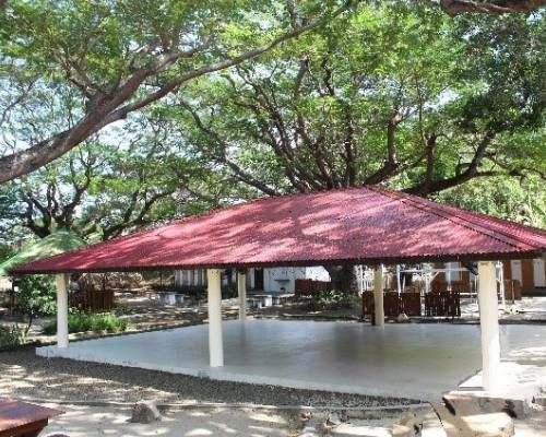 Red-roofed outdoor pavilion with white pillars and concrete floor, surrounded by trees.
