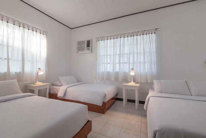 Bedroom with three beds, white linens, two windows with curtains, and bedside lamps.