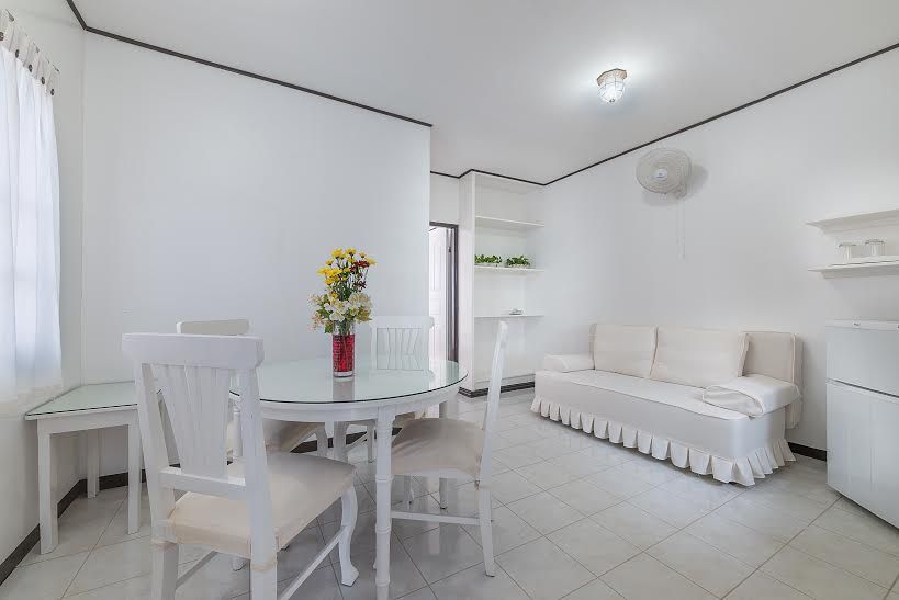 White living room with dining area, sofa, and refrigerator. A vase of flowers sits on the round table.