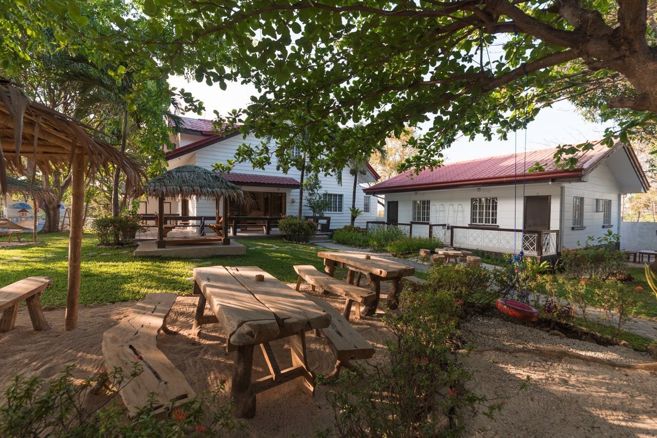 Outdoor picnic area with wooden tables, a gazebo, and two buildings under trees.