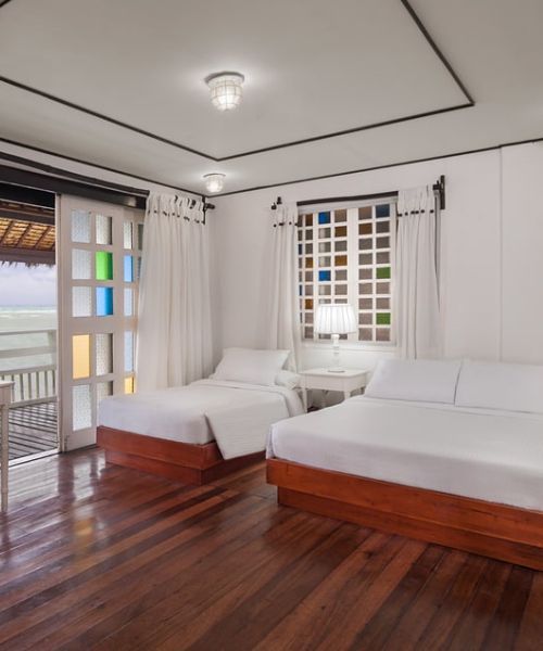 Bedroom with two beds, white walls, wooden floor. Balcony with sea view, colorful window panes.