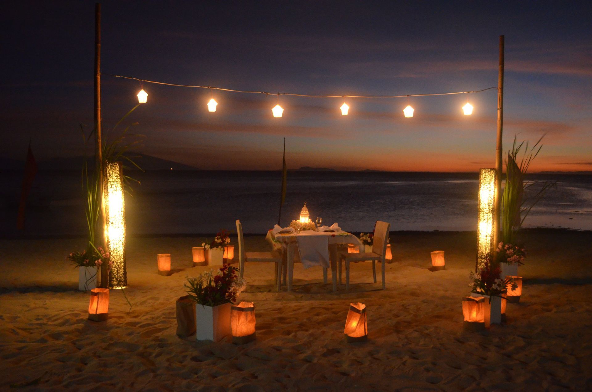 Romantic beach dinner setting with lanterns, string lights, and sunset.