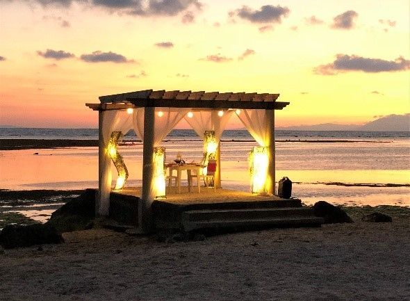 Gazebo with draped curtains and fairy lights on a beach at sunset. Table set for a meal.