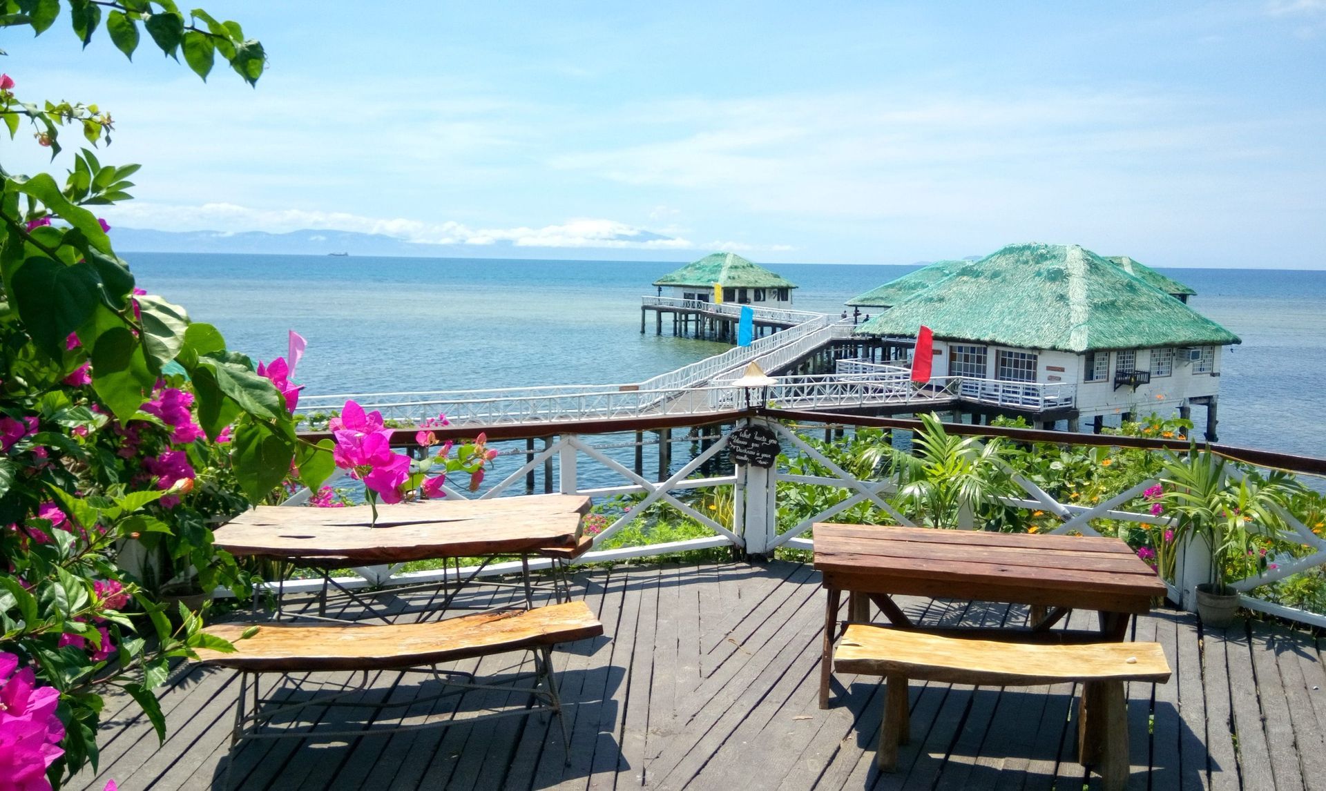 Wooden deck overlooking ocean with tables, flowers, and buildings on stilts with green roofs.