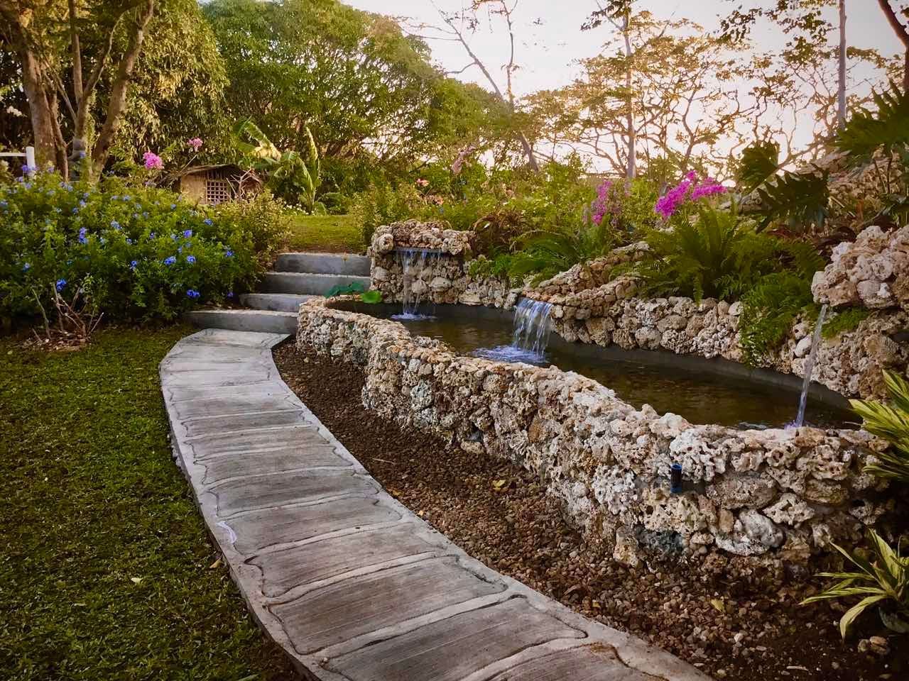 Stone pathway winds past a pond with waterfalls, lush greenery, and blooming flowers in a garden.