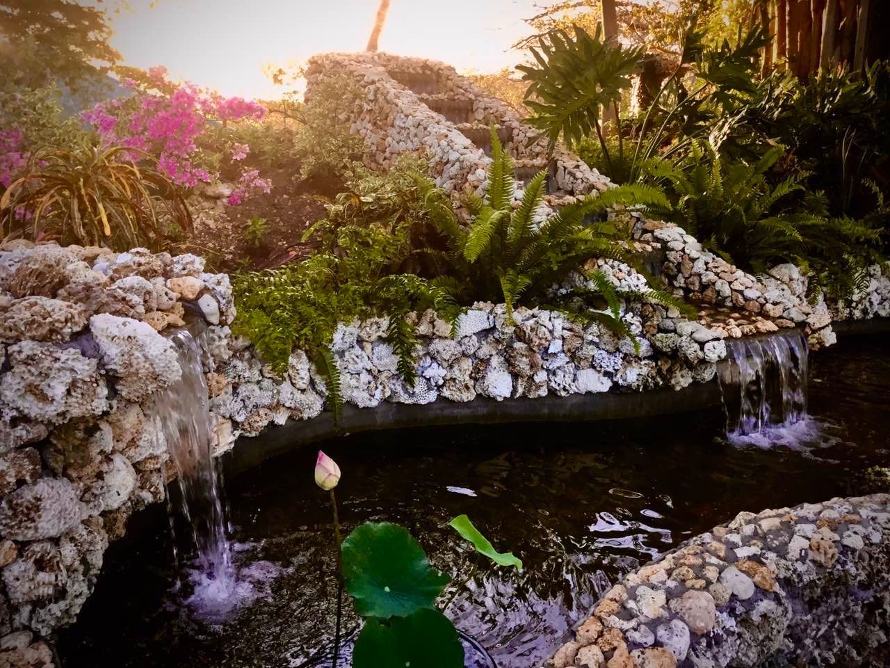Waterfalls cascading into a pond in a lush garden with stone edges, flowers, and greenery.
