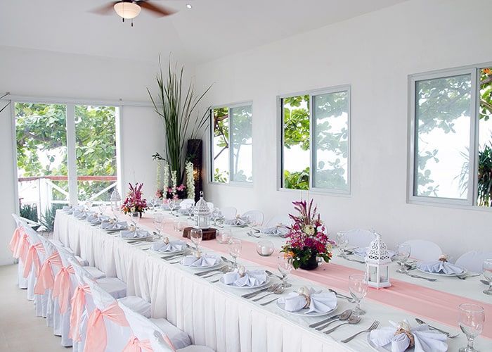 Long rectangular table set for a formal event, with pink runner and floral centerpieces; white chairs with pink bows.