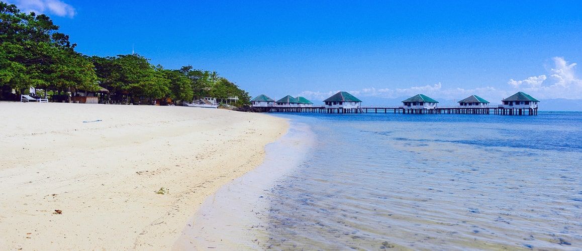Beach with white sand, clear blue water, and huts over the ocean under a bright blue sky.
