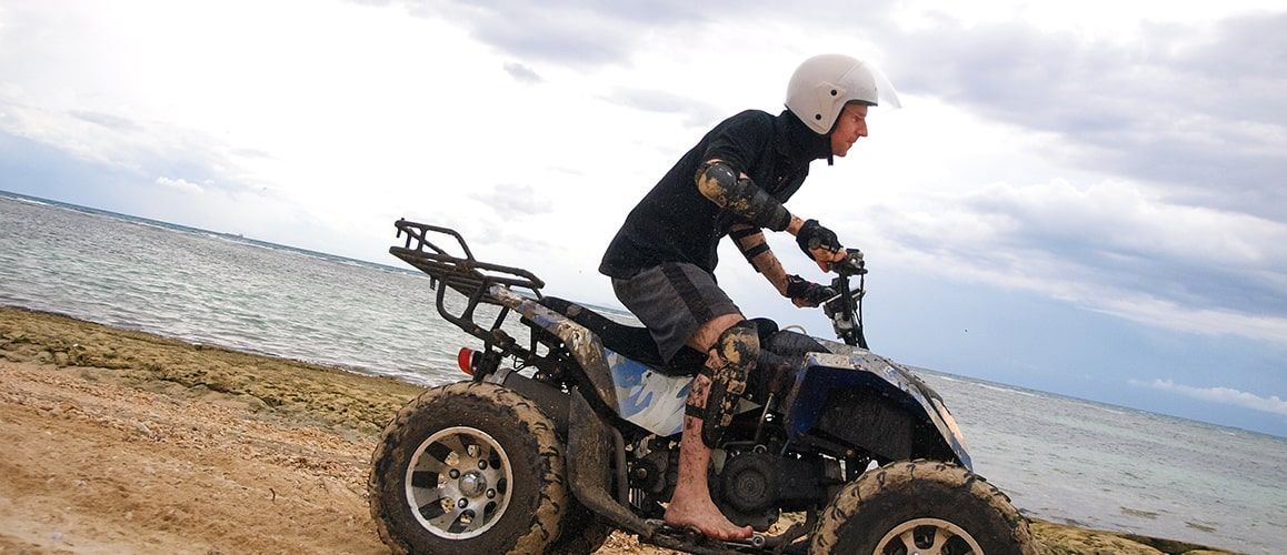 Person rides an ATV on a sandy beach near water. They wear a helmet and protective gear. Cloudy sky.